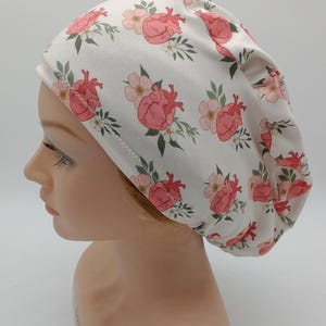 May include: White scrub cap with a repeating pattern of pink anatomical hearts and small pink flowers with green leaves. The cap is on a mannequin head.