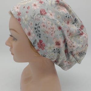 May include: A floral print scrub cap in a light gray color, adorned with pink and white flowers and green leaves. The cap is designed to cover the head and hair, commonly used in medical settings.