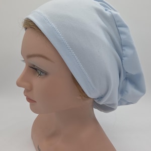 Puede incluir: Un gorro de tela azul claro con la parte trasera fruncida y una costura cosida a lo largo del borde delantero. El gorro se muestra en una cabeza de maniquí sobre un fondo blanco. Este artículo es adecuado para la protección del cabello o la moda.