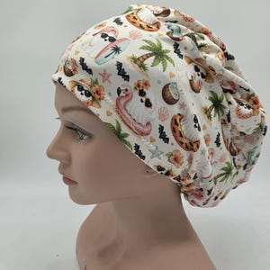 May include: A scrub cap featuring an off-white background with a repeating pattern of ghosts wearing sunglasses, tropical palm trees, pink flamingo pool floats, coconuts, seashells, hibiscus flowers, and small black bats scattered throughout.