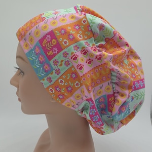 May include: A scrub cap featuring a vibrant patchwork pattern of floral designs in shades of orange, pink, light blue, and white. The fabric is gathered at the back with an elastic band, creating a rounded, voluminous shape that covers the head.