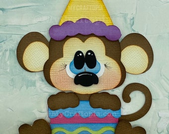 May include: A brown monkey wearing a purple and yellow party hat sits on a pink, blue, and green striped egg. The monkey has blue eyes and a black nose. The text "My Craftopia Created by Leslie Foley" is at the bottom of the image.