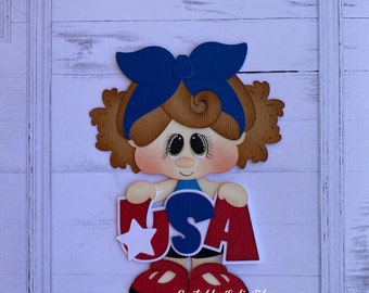 May include: A paper craft of a girl with brown hair wearing a blue bow holding a red, white, and blue USA sign. The sign has a white star on the left side.