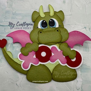 May include: A green paper dragon with pink wings and a red heart on its tail. The dragon holds a pink and red "XOXO" sign. The image includes the text "My Craftopia by Leslie Foley" and "@MyCraftopia.com".