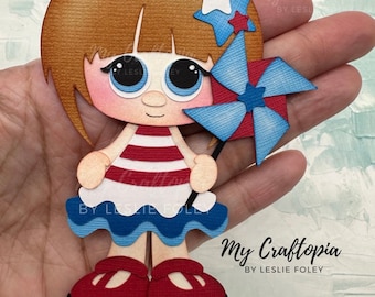 May include: A paper craft cutout of a girl wearing a red and white striped dress and holding a blue and red pinwheel. The girl has brown hair and blue eyes. The pinwheel has a white star on it. The text "My Craftopia by Leslie Foley" is at the bottom of the image.