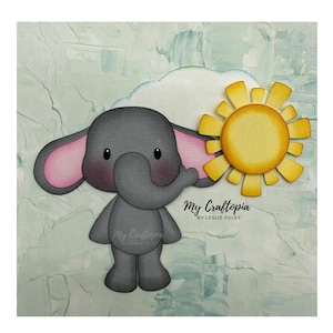 May include: A gray elephant with pink ears holding a yellow sun with a white cloud behind it. The text "My Craftopia by Leslie Foley" is below the elephant and sun.