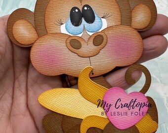 May include: A layered papercraft monkey holding a banana and a pink heart. The monkey has brown fur, blue eyes, and pink cheeks. The banana is yellow and brown. The heart has the text "My Craftopia by Leslie Foley". The image also includes the text "@MyCraftopia.com".
