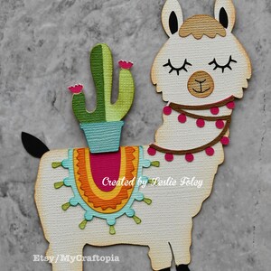 May include: A white llama with a brown head and black ears, wearing a pink and brown beaded necklace. The llama is carrying a green cactus in a blue pot on its back. The llama has a colorful saddle with a pink, orange, yellow, and blue design. The text "Created by Leslie Foley" and "Etsy/MyCraftopia" is printed on the llama.