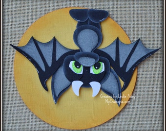 May include: A black paper bat with green eyes and white fangs hangs upside down against a yellow circle. The text "Created by Leslie Foley MyCraftopia.com" is below the bat.