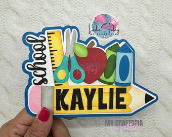 May include: A school-themed die-cut design featuring a pencil shape with the name "KAYLIE" in bold black letters. The design includes a ruler, scissors, apple, and crayons. The word "school" is written in a stylized font. The design is outlined in blue.