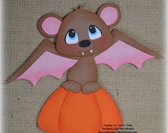 May include: A brown cartoon bat with pink wings and ears sits atop an orange pumpkin. The bat has blue eyes and a small smile. The image is a Halloween-themed craft.