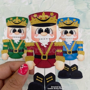 May include: Three papercraft nutcracker ornaments with gold glitter accents. The center nutcracker is red, white, and gold. The nutcracker on the left is green, white, and gold. The nutcracker on the right is blue, white, and gold. The ornaments are created by Leslie Arely Foley @MyCraftopia.com