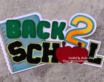May include: Back to school themed paper craft with the words "Back 2 School" in green, yellow, and black letters. Two red apples are below the words. The craft is on a white background.