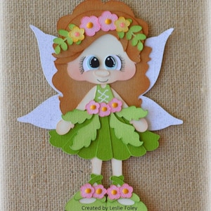 May include: A paper fairy with brown hair, large blue eyes, and white wings. The fairy wears a green dress with pink flower accents and green shoes with pink flowers. A floral crown adorns the fairy's head. Text at the bottom reads "Created by Leslie Foley".