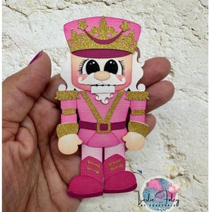 May include: A pink and gold paper cutout of a nutcracker soldier. The nutcracker has a pink uniform with gold accents, a gold crown, and white hair. The nutcracker is held in a hand against a textured background.