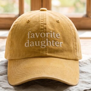 May include: Mustard yellow baseball cap with the embroidered text "favorite daughter" in white. The hat is made of a soft material and has a curved brim. It is sitting on a gray linen napkin on a wooden table.