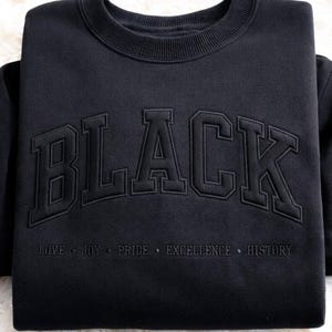 May include: Black sweatshirt with the word "BLACK" in large, raised lettering across the chest. Below the word "BLACK" are the words "LOVE • JOY • PRIDE • EXCELLENCE • HISTORY" in smaller lettering. The sweatshirt is folded on a white, fluffy surface.