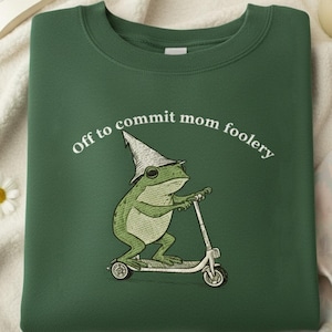 May include: Green sweatshirt with the phrase "Off to commit mom foolery" in white. The design features a green frog wearing a wizard hat and riding a scooter. The sweatshirt is on a white surface with a book, flowers, and a candle.