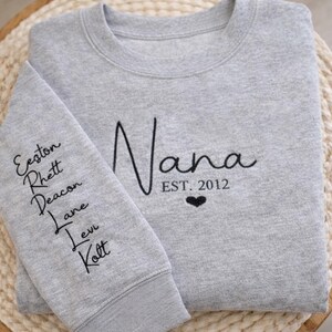 May include: A light gray sweatshirt with the word "Nana" in black script, "EST. 2012", and a heart. The left sleeve has embroidered names: Eeston, Rhett, Deacon, Lane, Levi, and Kolt. The sweatshirt is folded on a woven surface.