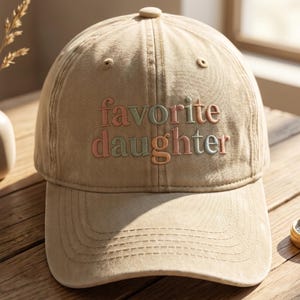 May include: A tan baseball cap with the embroidered text "favorite daughter" in pastel colors. The cap has a curved brim and a button on top. The hat is made of a soft, durable fabric.