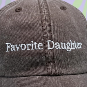 May include: A brown baseball cap with the embroidered text "Favorite Daughter" in white. The cap has a curved peak and a fabric strap closure. The cap is made of a durable, textured fabric.