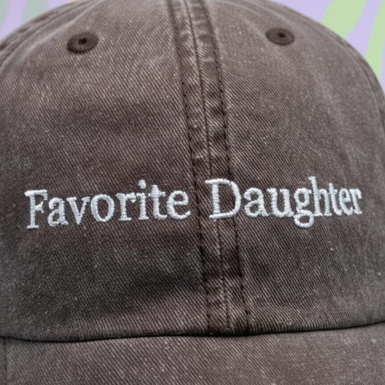 May include: A brown baseball cap with the embroidered text "Favorite Daughter" in white. The cap has a curved peak and a fabric strap closure. The cap is made of a durable, textured fabric.
