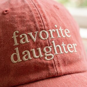 May include: A red baseball cap with the embroidered text "favorite daughter" in a cream-colored thread. The cap has a curved brim and a fabric strap closure.