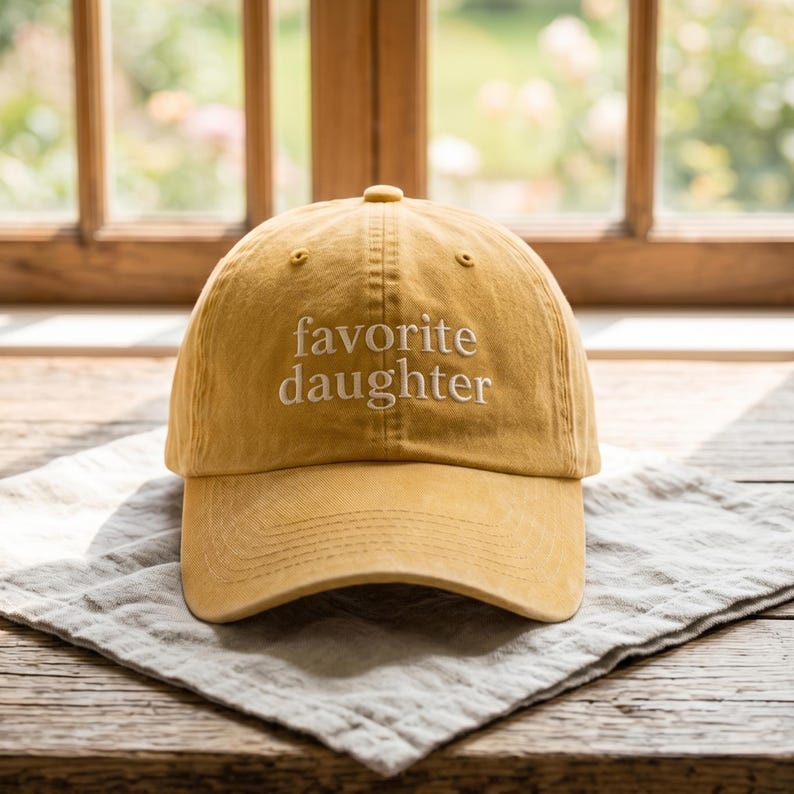 May include: Mustard yellow baseball cap with the embroidered text "favorite daughter" in white. The hat is made of a soft material and has a curved brim. It is sitting on a gray linen napkin on a wooden table.