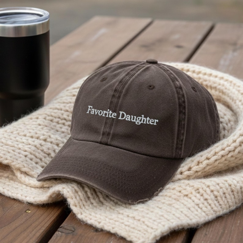 May include: A brown baseball cap with the text "Favorite Daughter" embroidered in white. The cap has a distressed look and is placed on a cream knitted scarf. A black tumbler is in the background, on a wooden surface.