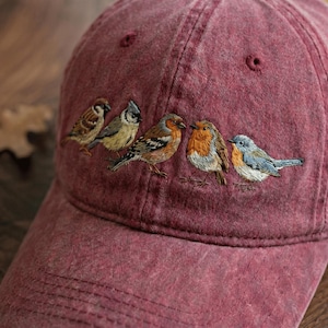 May include: A maroon baseball cap with an embroidered design of five colorful birds. The birds are detailed with various shades of brown, orange, blue, and gray. The cap is made of a soft, textured fabric.