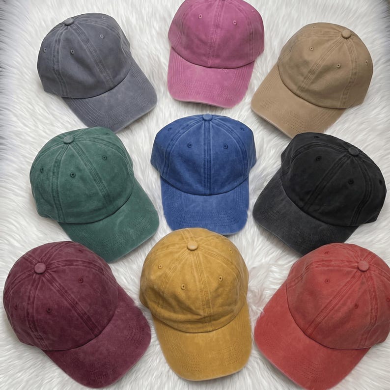 May include: A collection of nine baseball caps in various colors, including gray, pink, tan, green, blue, black, burgundy, yellow, and red. The caps are arranged on a white, fluffy surface.
