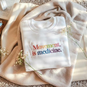 Pu&ograve; includere: Una felpa bianca con la scritta ricamata "Movement is medicine" nei colori dell'arcobaleno. La felpa &egrave; piegata su una coperta beige, con piccoli fiori bianchi e un libro sullo sfondo.