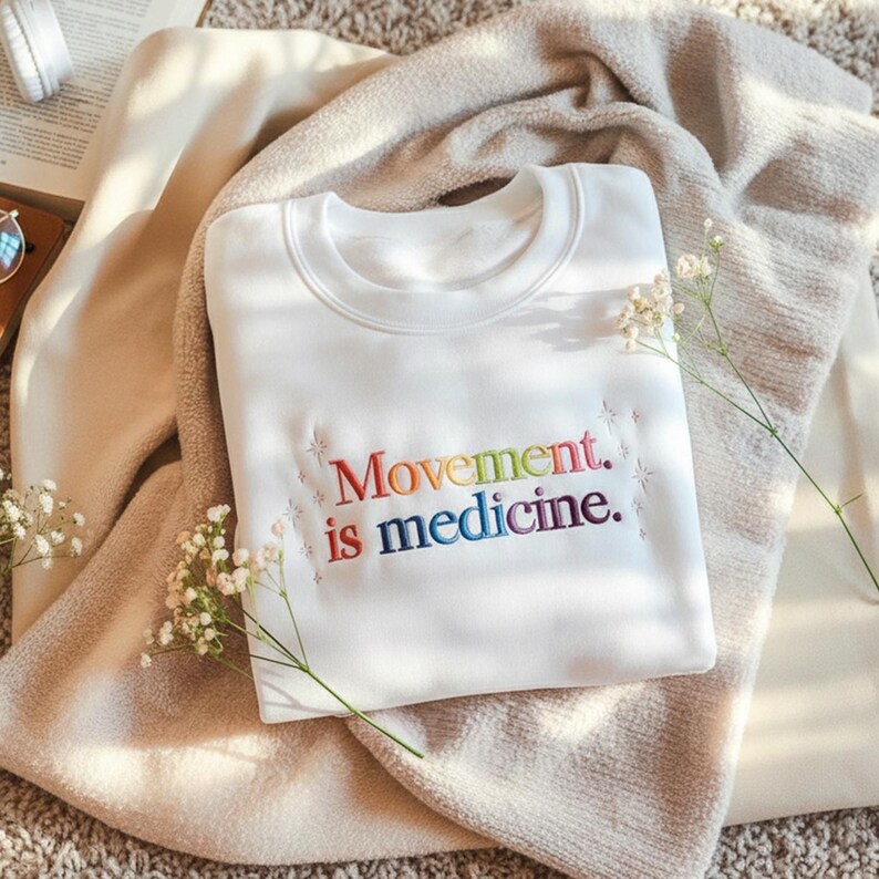 Pu&ograve; includere: Una felpa bianca con la scritta ricamata "Movement is medicine" nei colori dell'arcobaleno. La felpa &egrave; piegata su una coperta beige, con piccoli fiori bianchi e un libro sullo sfondo.