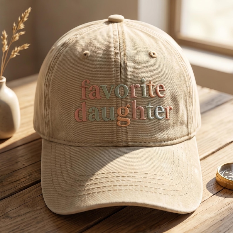 May include: A tan baseball cap with the embroidered text "favorite daughter" in pastel colors. The cap has a curved brim and a button on top. The hat is made of a soft, durable fabric.