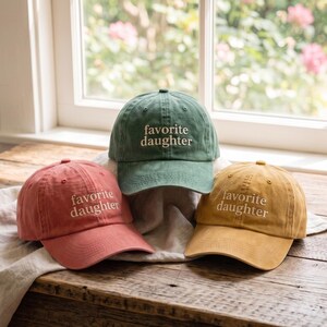 May include: Three baseball caps in shades of red, green, and yellow. Each cap has the embroidered text "favorite daughter" in white. The caps are displayed on a wooden surface near a window.