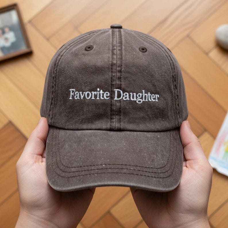 May include: A brown baseball cap with the embroidered text "Favorite Daughter" in white. The cap has a curved peak and a classic six-panel design. The hat is made of a soft, worn material.
