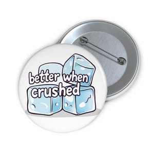 Op de afbeelding: Een witte button met de tekst "better when crushed" boven een stapel lichtblauwe cartoon ijsblokjes. Op de achtergrond is een tweede button zichtbaar, met een zilveren speld.