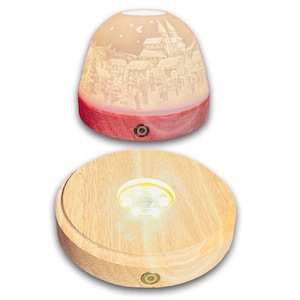 Könnte beinhalten: Eine dekorative Lampe mit einer detaillierten Winterdorf-Szene, die auf der oberen Kuppel geätzt ist. Die Kuppel sitzt auf einem roten Sockel. Die Lampe steht auf einem runden Holzsockel mit einem zentralen Licht.