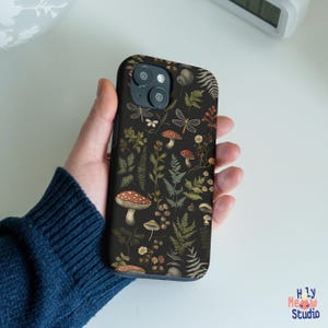 Könnte beinhalten: Schwarze Handyhülle mit einem Blumenmuster mit Pilzen, Farnen und Schmetterlingen. Die Hülle hat einen dunklen Hintergrund und eine Vielzahl von Farben, darunter Rot, Grün und Braun.
