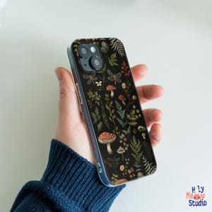 May include: A black phone case featuring a botanical design with mushrooms, ferns, and dragonflies. The case has a clear border and a glossy finish. The design includes red, white, and green accents.