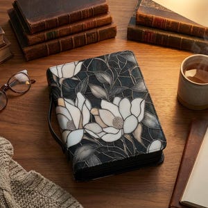 May include: A book cover with a floral design, showing magnolia flowers in cream and white on a dark backdrop. It sits on a wooden surface with books, a mug of tea, spectacles, and a notebook.
