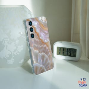 May include: A smartphone case with a pearlescent, agate-like design in shades of peach, cream, and white. The case has three camera lenses and a clear border. The image also includes a white digital clock and a decorative vase.