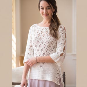 Peut inclure: Un haut blanc en crochet avec un motif floral et des manches trois-quarts. Le haut présente des détails découpés sur les manches et une fente sur l'ourlet. La femme porte un bracelet et une jupe violet clair.