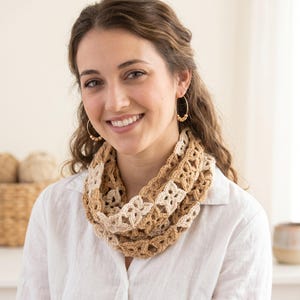 Peut inclure: Écharpe infinie en crochet beige et marron clair. Le motif ajouré présente un motif floral répété. L'écharpe est portée autour du cou. La femme porte une chemise blanche et des créoles dorées.