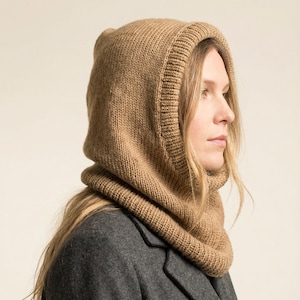 May include: A tan-colored knitted hood and cowl combination. The hood is designed to cover the head and neck, with a ribbed knit trim around the face and neck. The garment is made of a soft, warm material, perfect for cold weather. The model is wearing a gray coat.