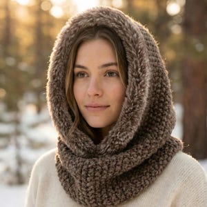 May include: A brown knit hood with a faux fur trim. The hood is worn over a cream-colored sweater. The image is taken outdoors with a blurred background of trees and sunlight.