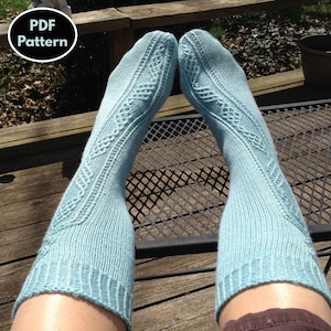 May include: A pair of light blue knitted socks with a cable pattern. The socks are worn on a person's legs and are sitting on a wooden surface with a metal grate.