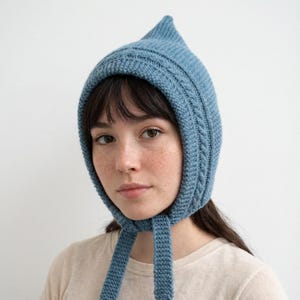 May include: A blue knitted hood with a pointed top and braided details. The hood has long ties that can be secured under the chin. The knitted hat is a soft, cozy accessory.