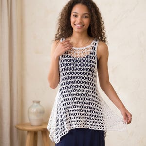 Peut inclure: Un haut en crochet blanc à tissage ouvert, avec un col rond et un ourlet légèrement asymétrique. Le haut sans manches semble être porté sur un vêtement bleu foncé. Le motif en crochet crée un design en forme de treillis.