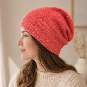 Puede incluir: Un gorro de punto de color coral vibrante con un borde acanalado y una superficie suave y texturizada. El gorro está confeccionado con un patrón de punto fino que proporciona un aspecto liso, cubriendo la cabeza con un ajuste holgado.